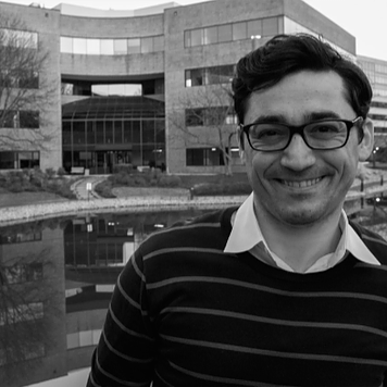 A black and white photograph of a smiling man wearing glasses in front of a modern building. No Description Available.